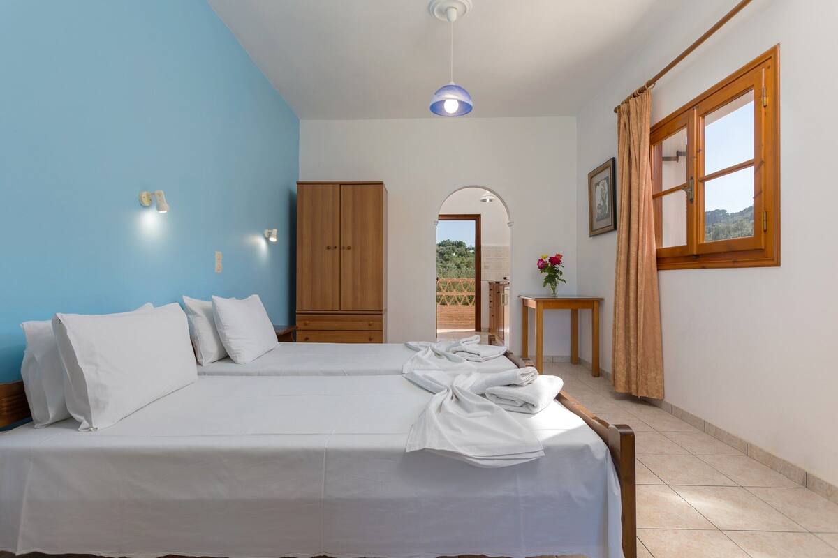 A bedroom with two beds, a wooden wardrobe, a small table with a vase, and a window with curtains.