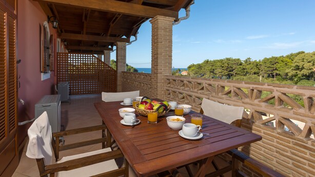 A wooden table set with breakfast items, including fruits, juice, and coffee, on a balcony with a view of the sea and greenery. - Anemomylos Studios (Zakynthos)