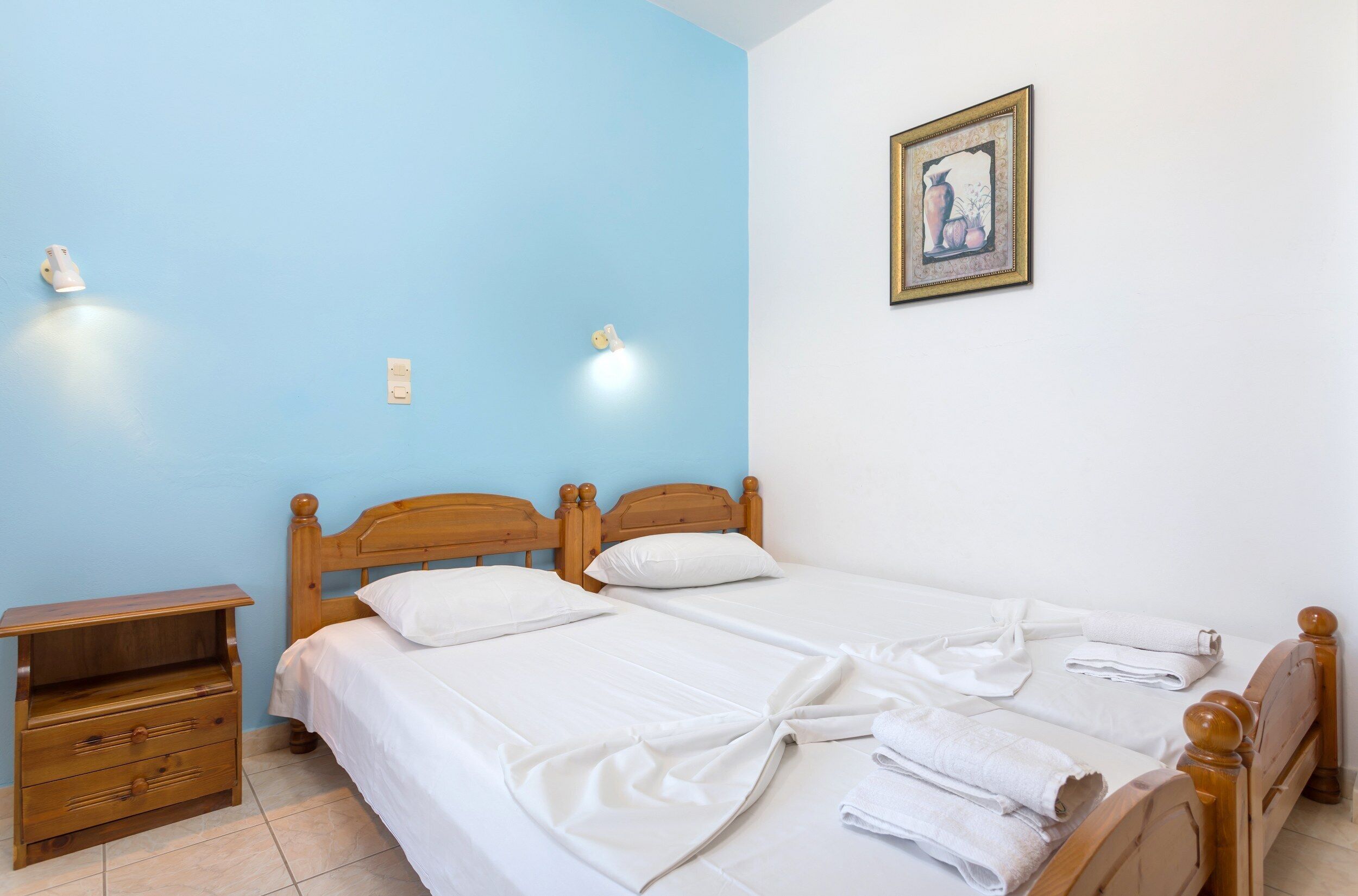 A double bed with white linens, a wooden nightstand with drawers, and a wall-mounted light fixture.