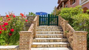 A set of stone steps leading to a building with a green gate, surrounded by red flowers and greenery. - Anemomylos Studios (Zakynthos)