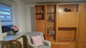 A living room with a grey armchair, a wooden cabinet with glass doors, a small table, and a window with blinds. - Ferienwohnung Anni (Dörfles-Esbach)