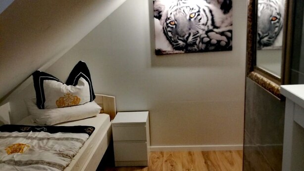 A bedroom with a sloped ceiling, a bed with a patterned bedspread, a bedside table, a nightstand, a round rug, and a wall-mounted tiger painting. - Ferienwohnung Anni (Dörfles-Esbach)