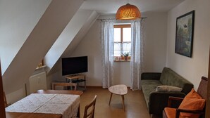 A cozy living room with a television, a sofa, a dining table, and a window with curtains. - Ferienwohnung Abel (Heideck)