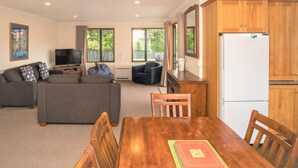A living room with a sofa, TV, and dining table with chairs. There is a kitchen area with wooden cabinets and a refrigerator. - Mountain Views from Meadowstone (Wānaka)