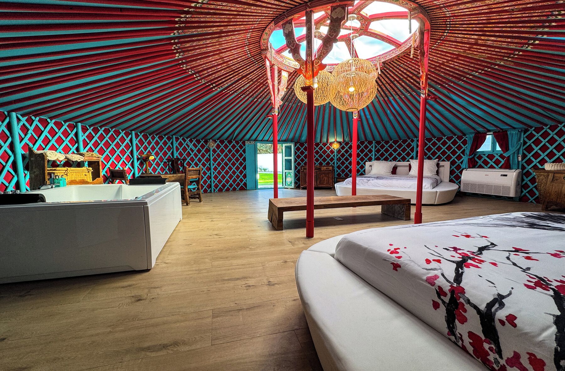 A spacious, well-lit tent with a central wooden structure, a large bed, a bench, and a small table with a lamp.