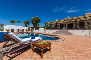 A poolside area with a lounge chair, a table with fruit and a drink, and a covered patio with plants.