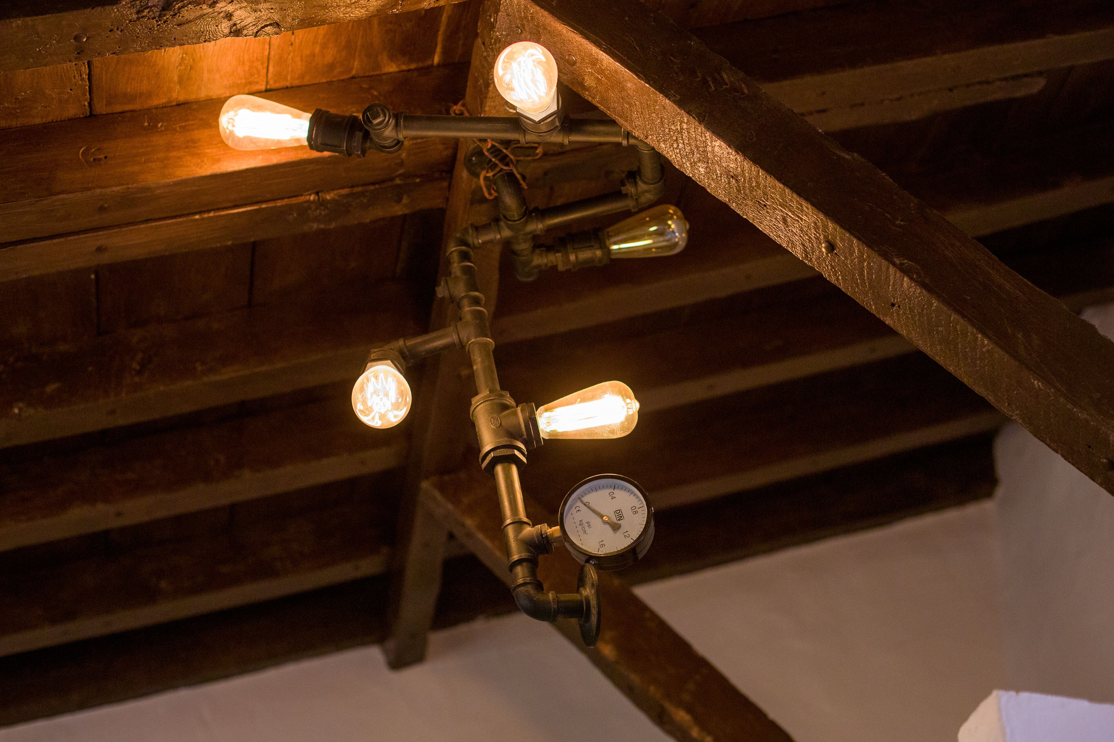 A ceiling with exposed wooden beams and a lighting fixture with exposed bulbs and a pressure gauge.