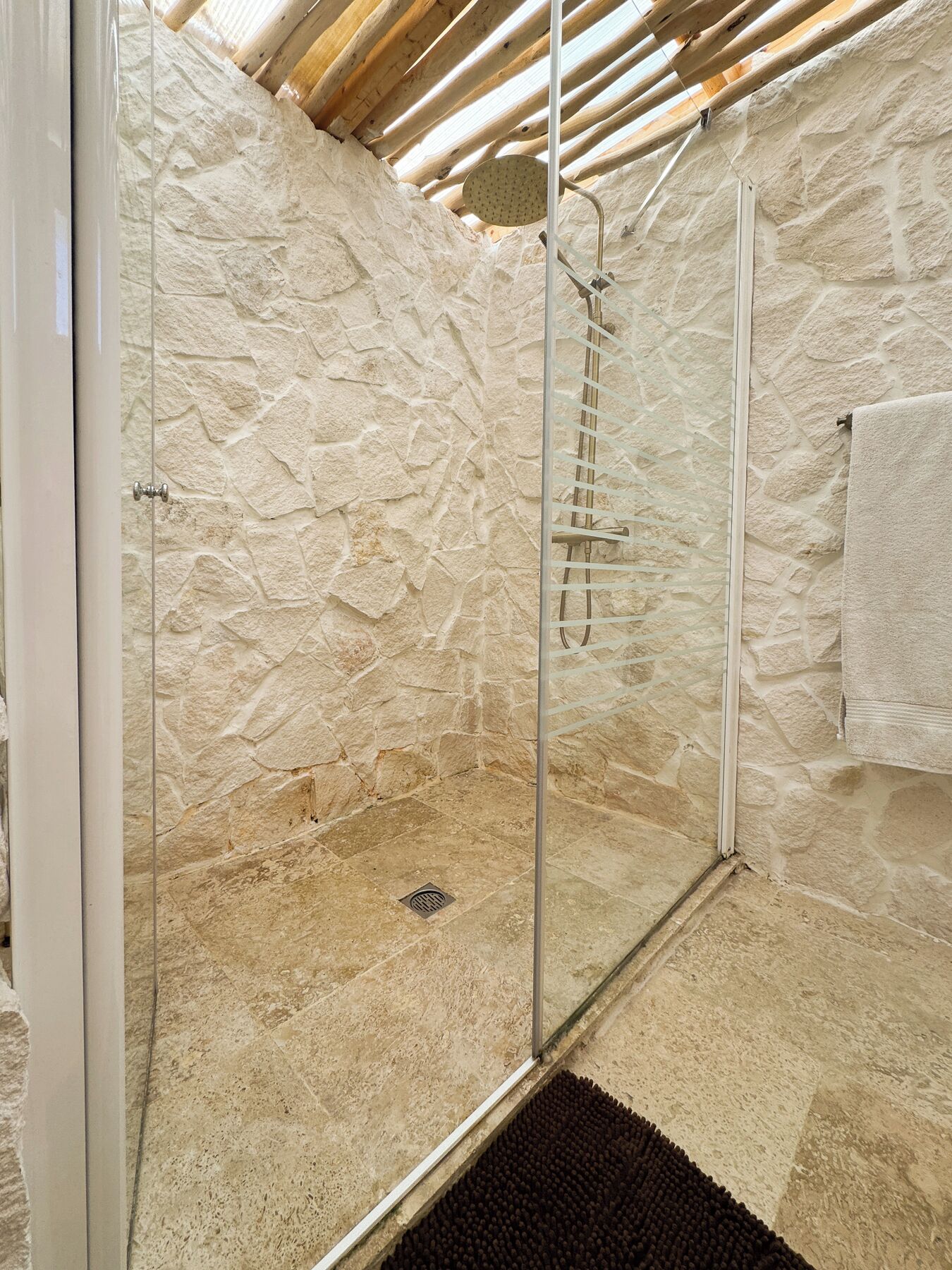 A glass-enclosed shower with a stone wall backdrop, a metal ladder, and a towel hanging on the wall.