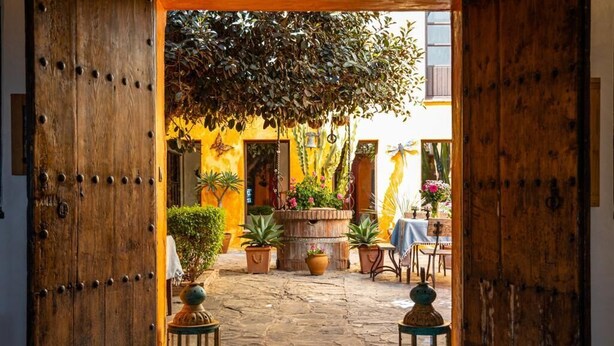 Une porte en bois ouverte donne sur une cour intérieure où se trouvent des plantes en pot, une table recouverte d’une nappe bleue et des lanternes.