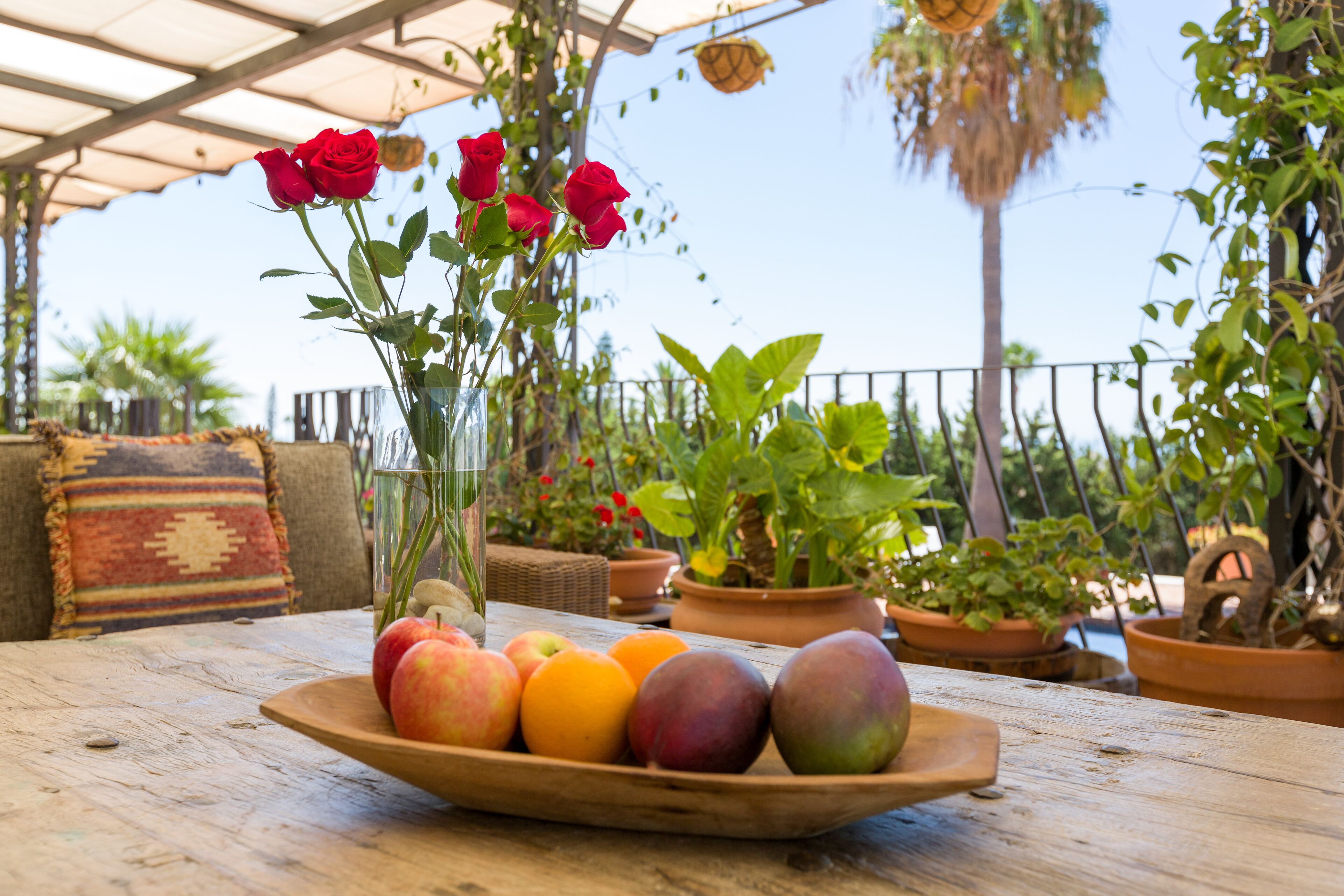 A wooden table with a fruit bowl containing apples, oranges, and peaches, and a vase with red roses.