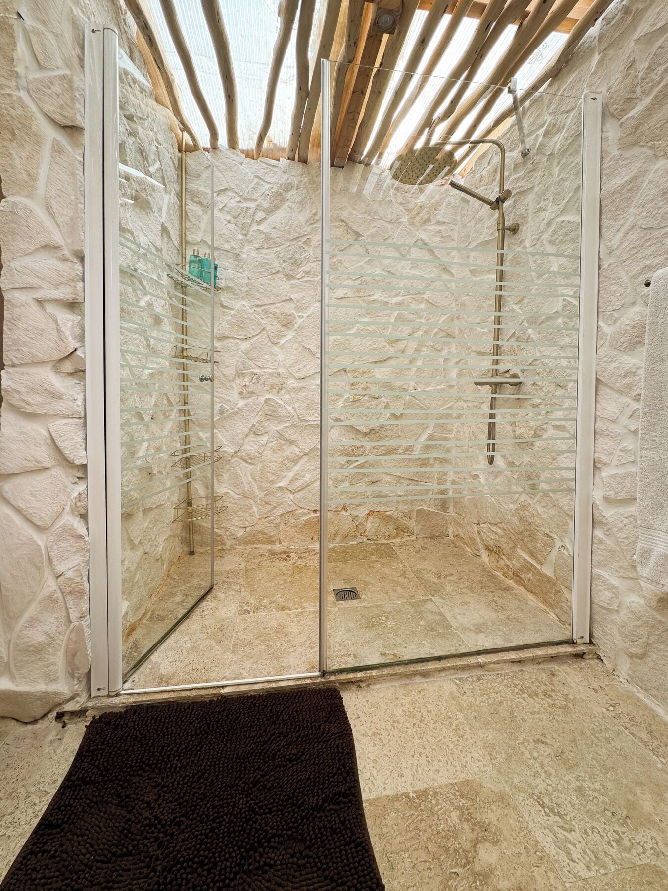Un espace de douche aux murs en pierre, doté de portes en verre, d’un pommeau de douche en métal et d’un tapis de bain noir.