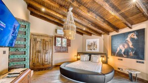 Une chambre avec un plafond en bois, un grand lit, un lustre, un tableau représentant un cheval et un téléviseur fixé au mur.