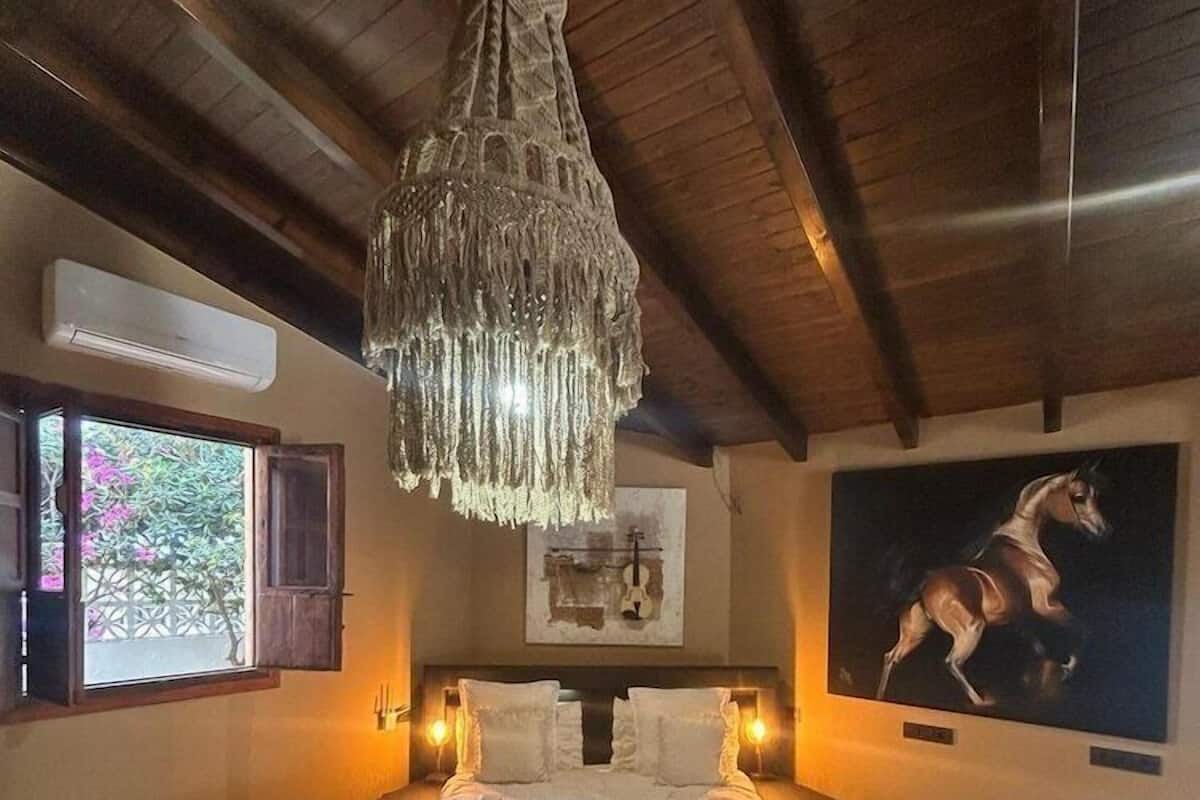 Une chambre à coucher avec un grand lit, un lustre, une fenêtre dont le volet est ouvert, et un tableau représentant un cheval.