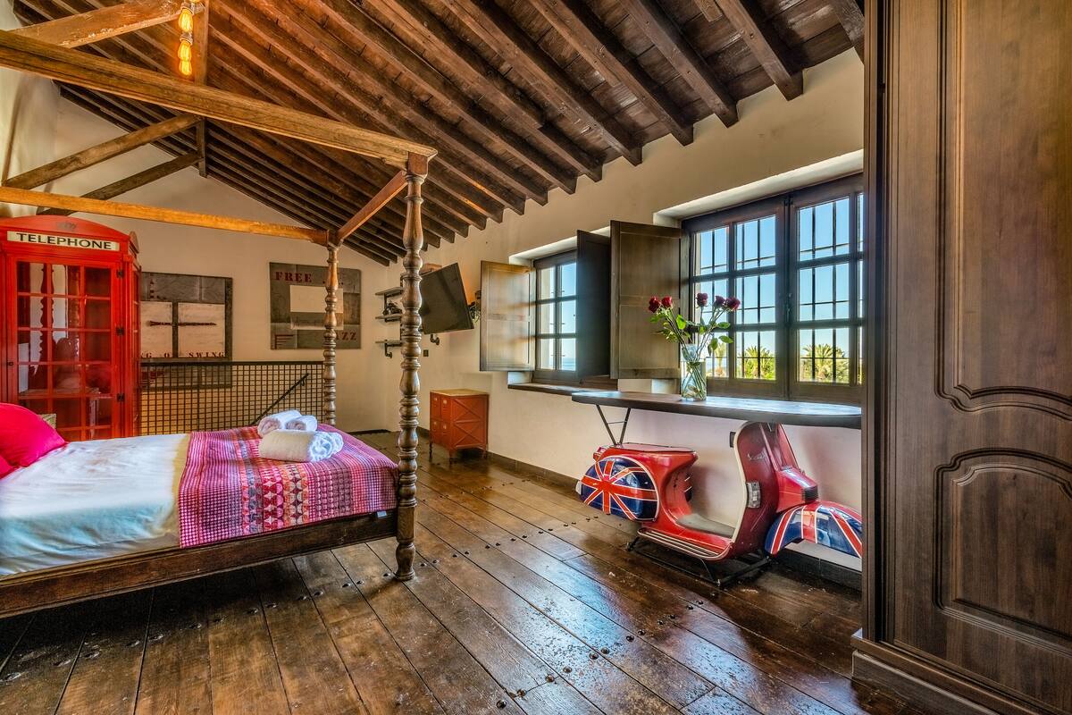 Une chambre dotée d’un lit à baldaquin, d’une cabine téléphonique rouge, d’un scooter vintage et d’une grande fenêtre donnant sur un paysage verdoyant.