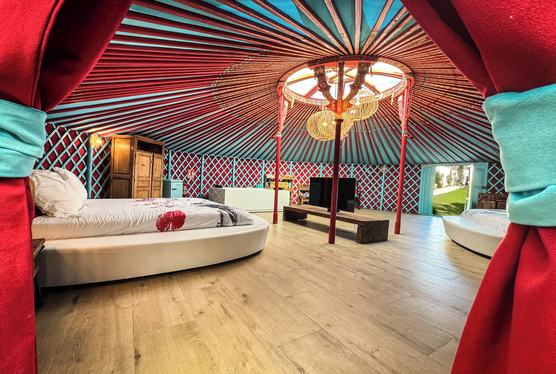 Une pièce circulaire et chaleureuse, dotée d’un plancher en bois, d’un lit, d’une télévision et d’une structure de plafond originale et ouverte, avec un luminaire suspendu.