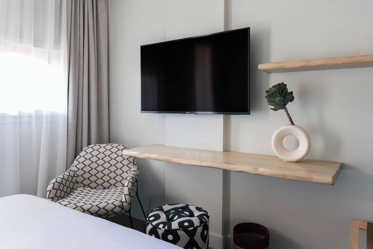 Habitació d'hotel moderna amb un llit, un otomà amb dibuixos, un petit reposapius, un televisor fixat a la paret i prestatges de fusta amb un objecte decoratiu.