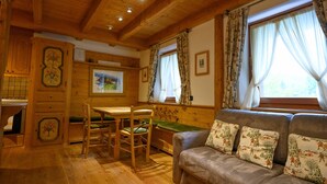 Interior acolhedor de uma cabana de madeira, com área de jantar, sofá com almofadas florais e janela com cortinas.