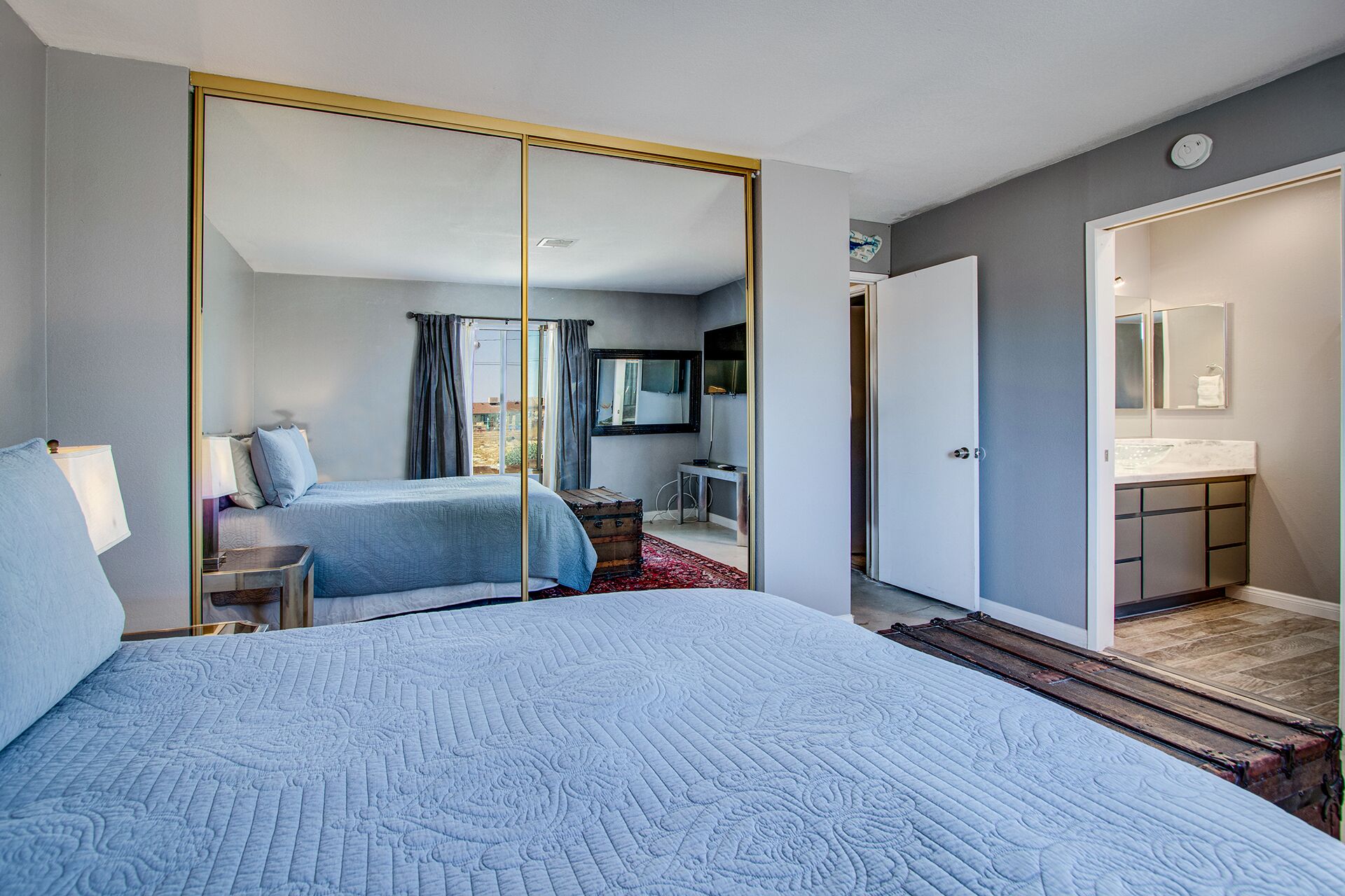 Un dormitorio con una cama grande, un armario con espejo, un escritorio y un baño con lavamanos y espejo.