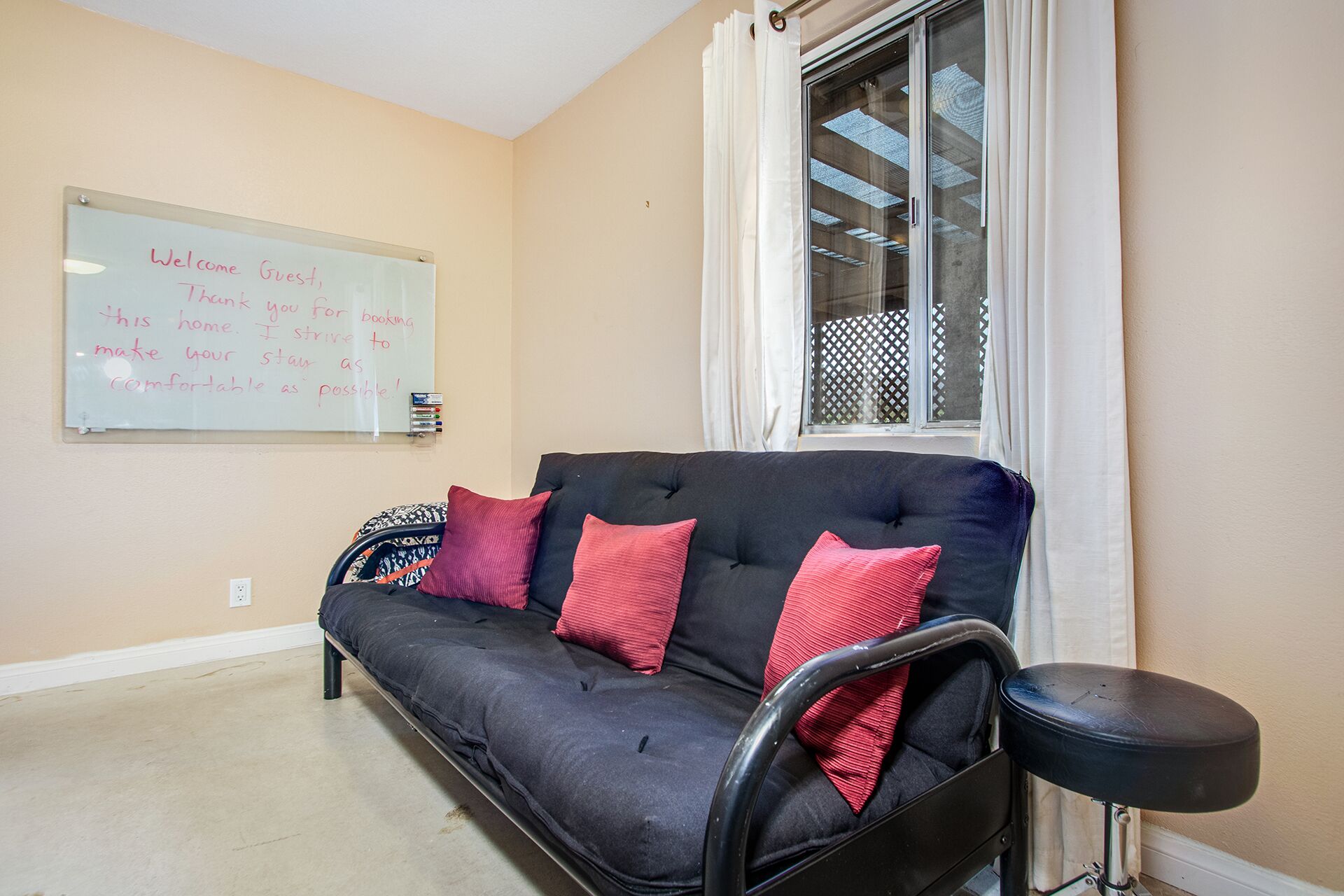 A room with a black sofa, red cushions, a whiteboard with a welcome message, a window with curtains, and a small table.