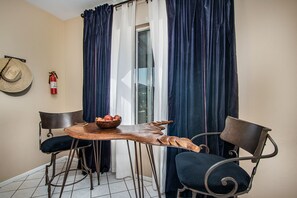 Un petit coin repas avec une table ronde, deux chaises, un saladier de fruits, un extincteur et un chapeau de paille accroché au mur.