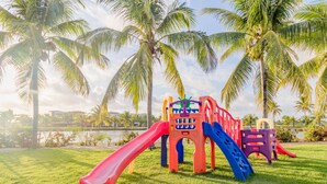 Un parque infantil con toboganes y estructuras para escalar, rodeado de palmeras y una masa de agua.