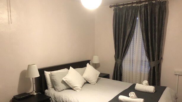 A neatly made bed with white linens and pillows, a bedside lamp, and a dark-colored blanket. - 1 Bed-sleeps 4-free Parking-15 Min to Putney (London)