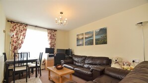 A living room with a brown leather sofa, a wooden coffee table, and a dining area with a table and chairs. There are three framed pictures on the wall, a chandelier, and a lamp. - 1 Bed-sleeps 4-free Parking-15 Min to Putney (London)