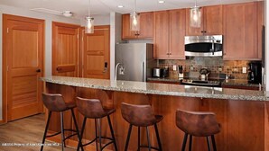 A modern kitchen with wooden cabinets, a stainless steel refrigerator, a microwave, and a gas stove. There are three bar stools and pendant lights above the counter.