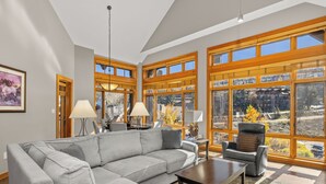 A spacious living room with a gray sectional sofa, a dark coffee table, and a leather armchair. Large windows offer a view of the outdoors.