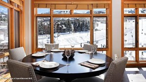 A dining area with a round table set for four, large windows with a snowy mountain view, and wooden paneled walls.