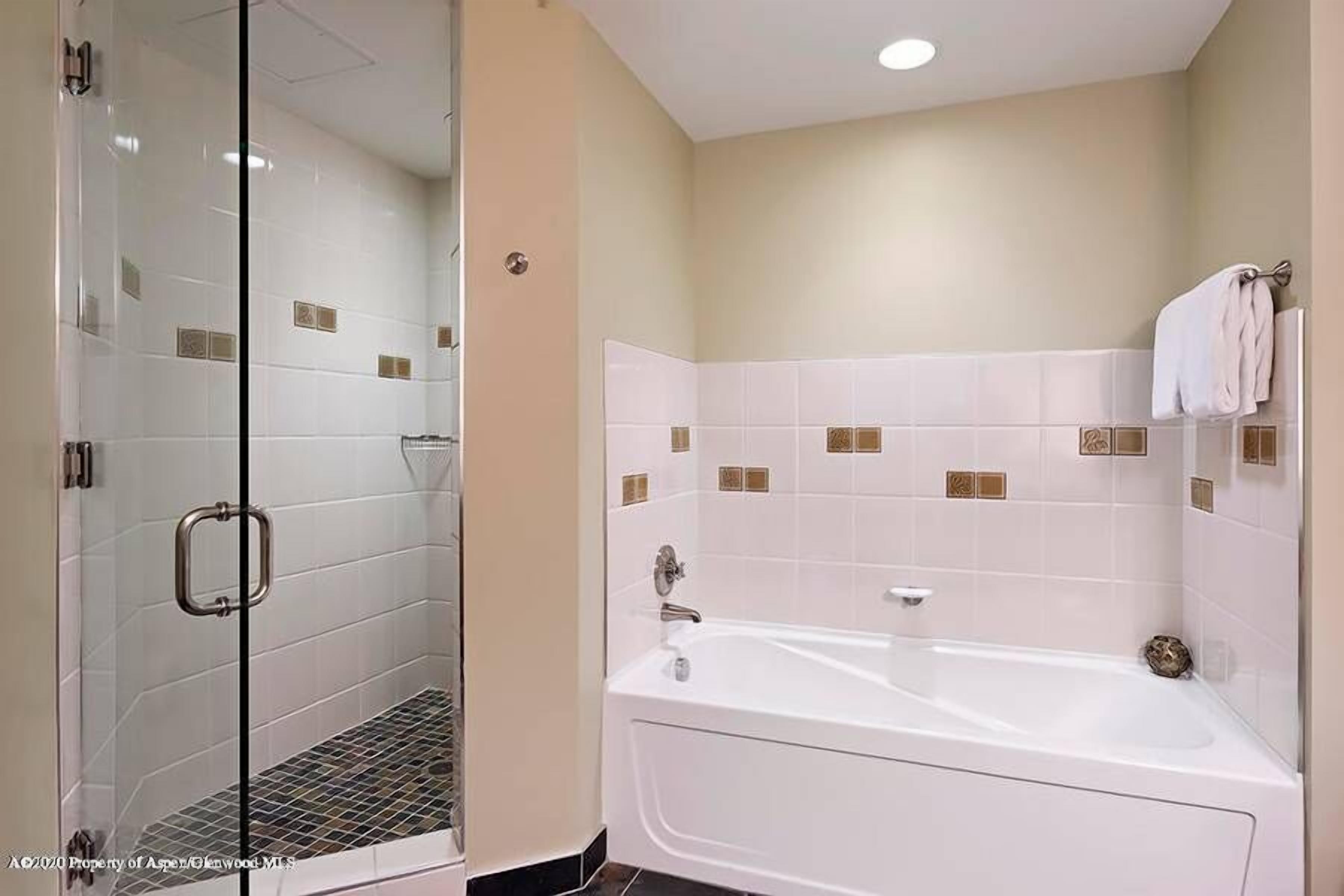 A modern bathroom with a glass-enclosed shower, a white bathtub, and beige tiled walls.