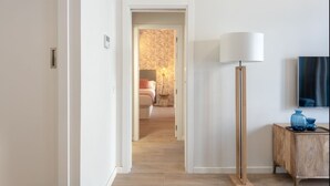 Un couloir mène à une chambre qui contient un lit, une table de chevet et une lampe. Un téléviseur est fixé au mur, et il y a un meuble en bois orné de vases bleus.