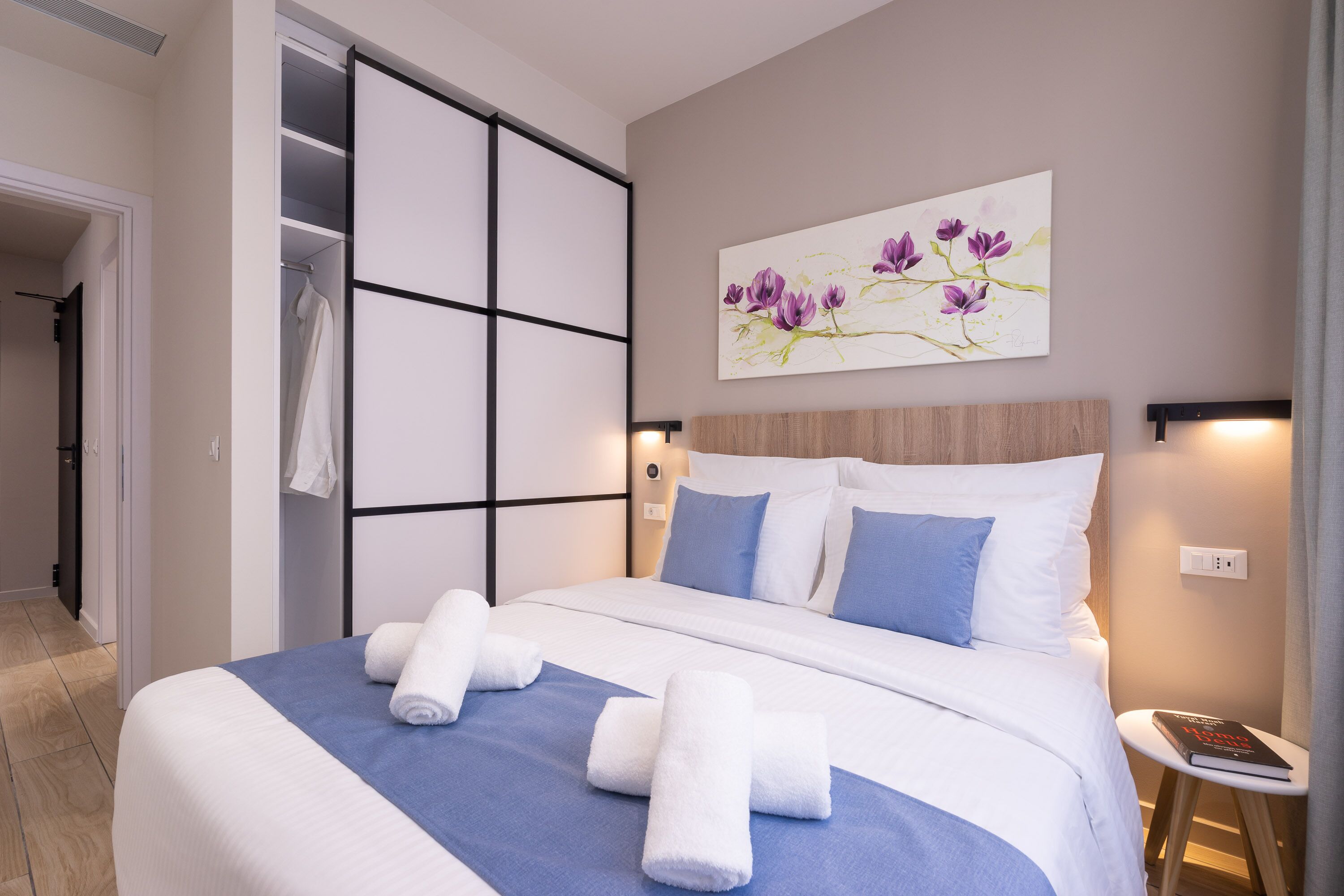 A neatly made bed with white and blue bedding, a wall-mounted artwork of purple flowers, and a bedside table with a book.