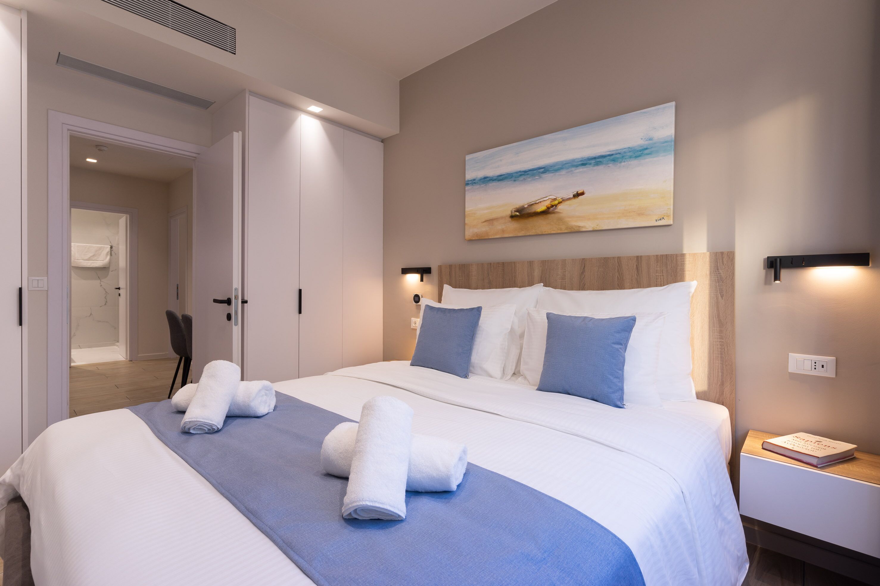 Une chambre d’hôtel avec un grand lit, un tableau représentant une scène de plage et une table de chevet avec un livre.