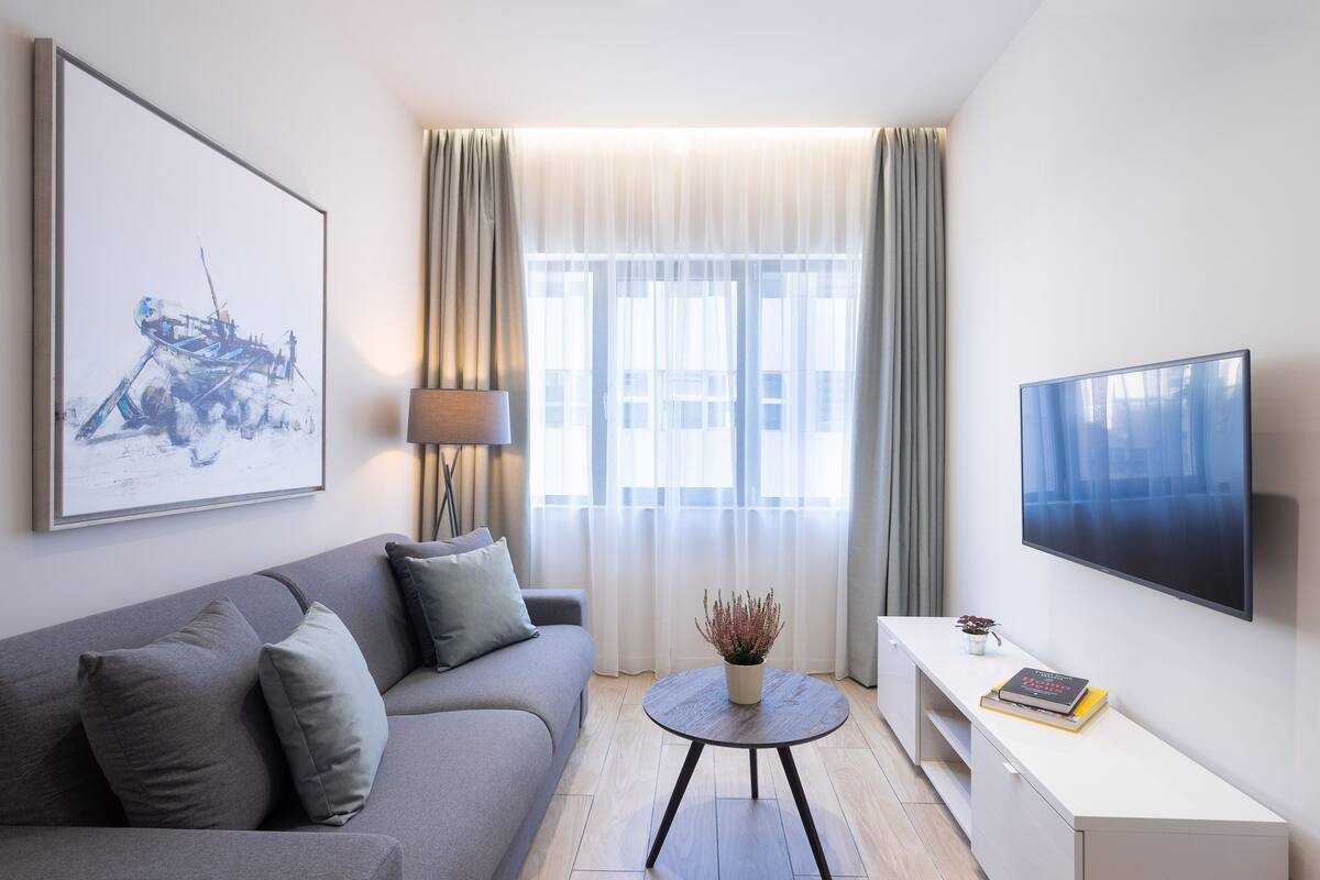 Un moderno salón con un sofá gris, una mesa de centro redonda con una planta, un televisor de pantalla plana y una obra de arte enmarcada en la pared.