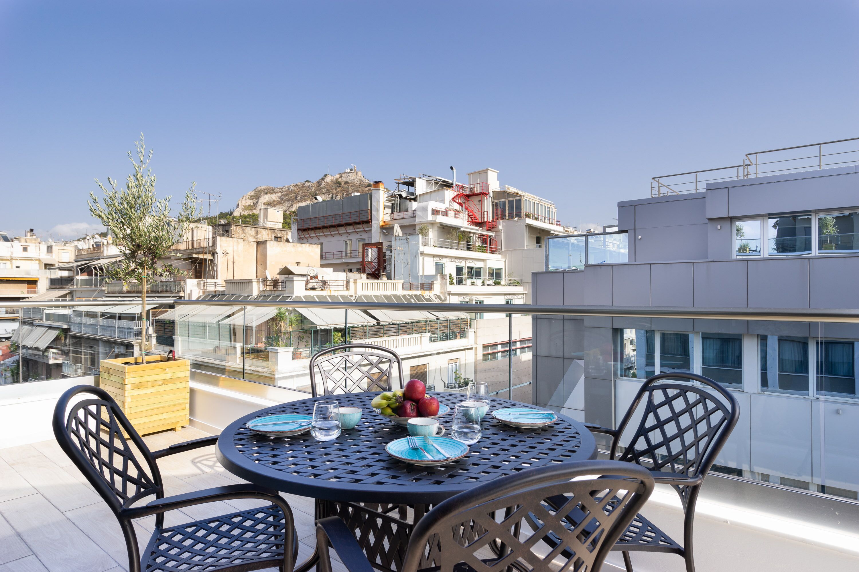 A rooftop terrace with a round table set for dining, surrounded by chairs, and a view of the cityscape in the background.
