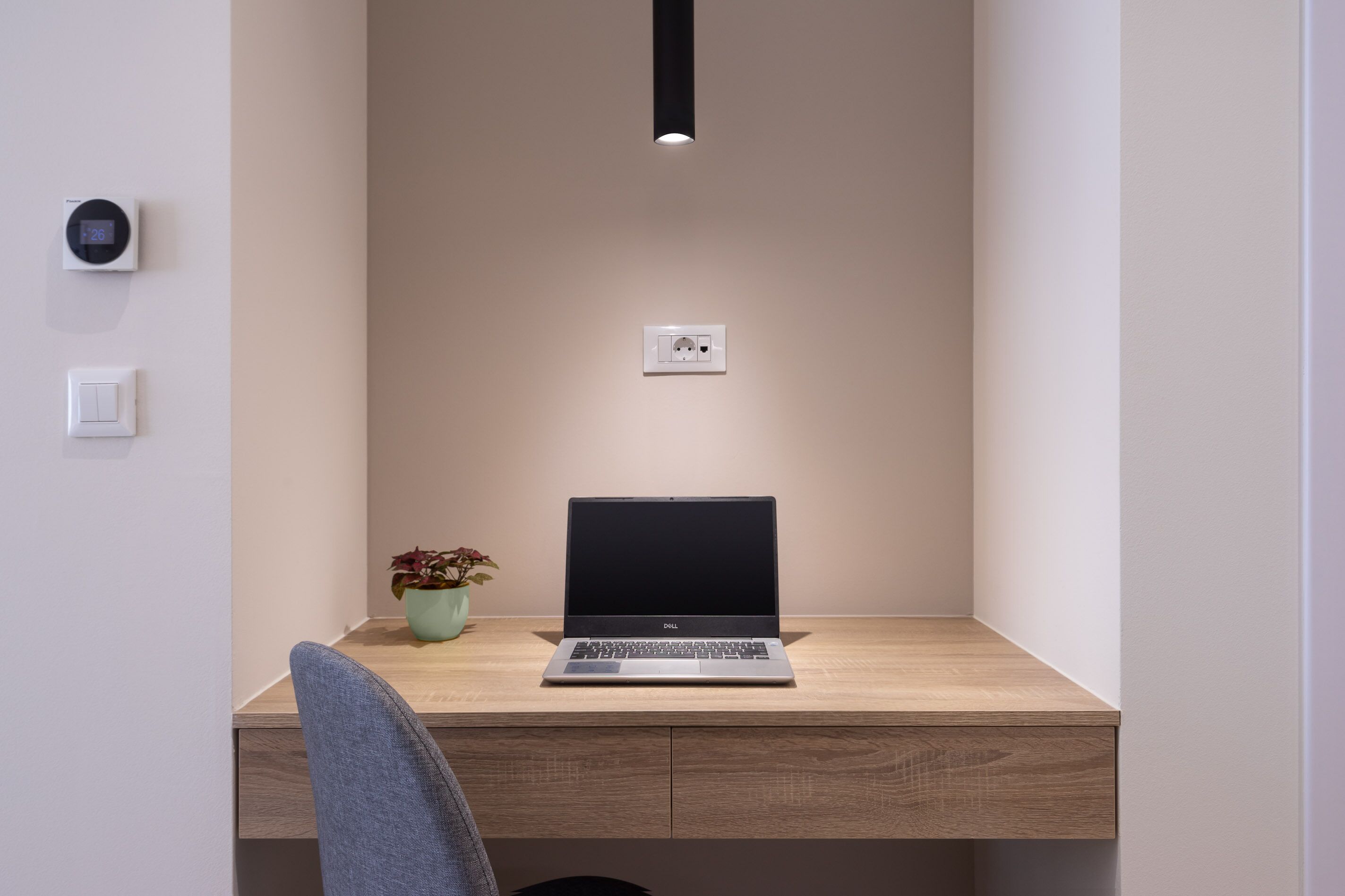 A minimalist workspace with a laptop on a wooden desk, a potted plant, and a wall-mounted thermostat.