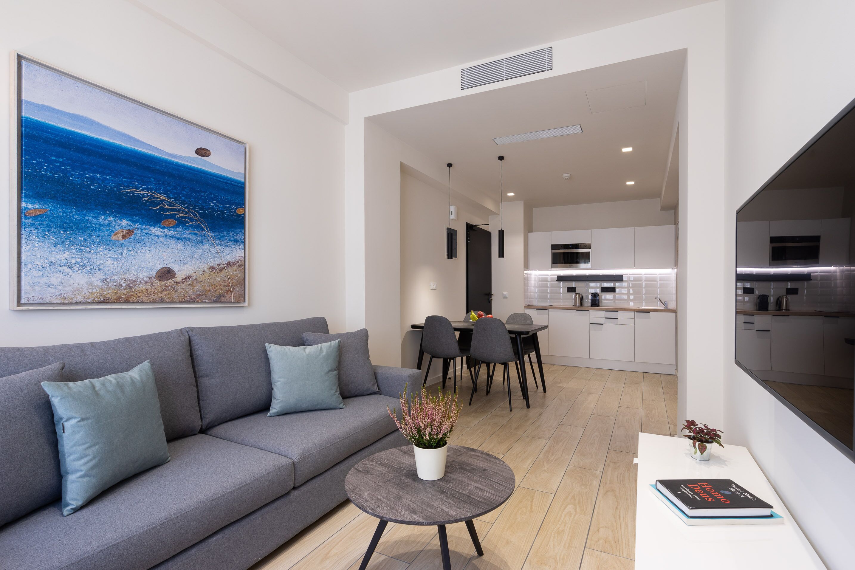 Ein modernes Wohnzimmer mit einem grauen Sofa, einem runden Couchtisch und einem Essbereich mit schwarzem Tisch und Stühlen. Im Hintergrund ist eine Küche zu sehen.