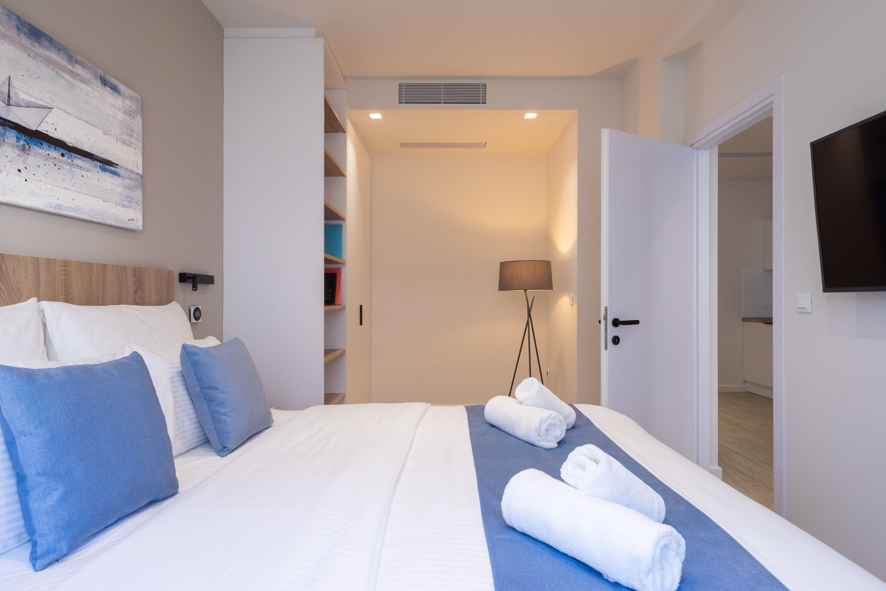 A hotel room with a bed, pillows, a blue towel, a lamp, and a TV.