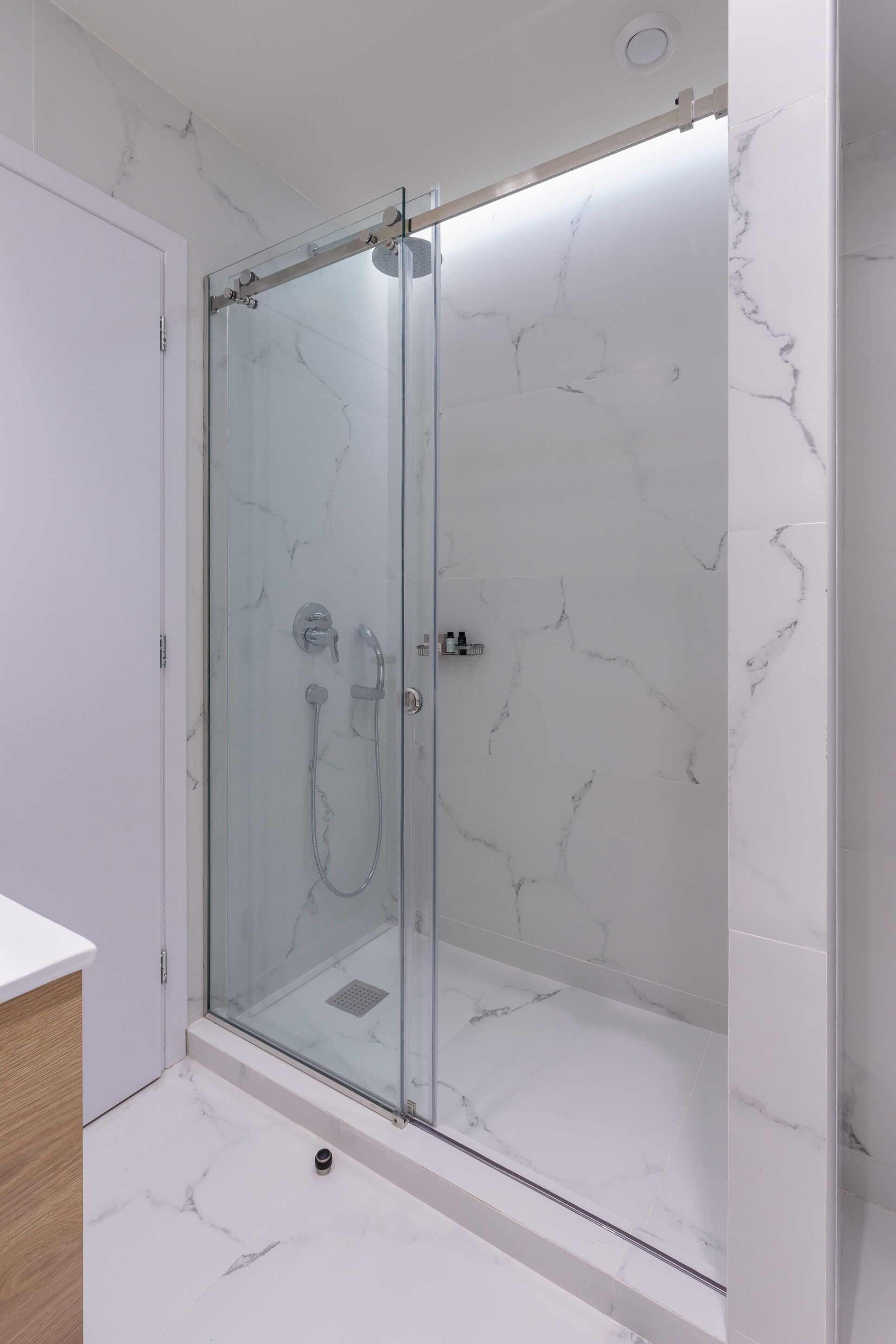 A glass-enclosed shower with a marble wall and floor, a white door, and a wooden cabinet.