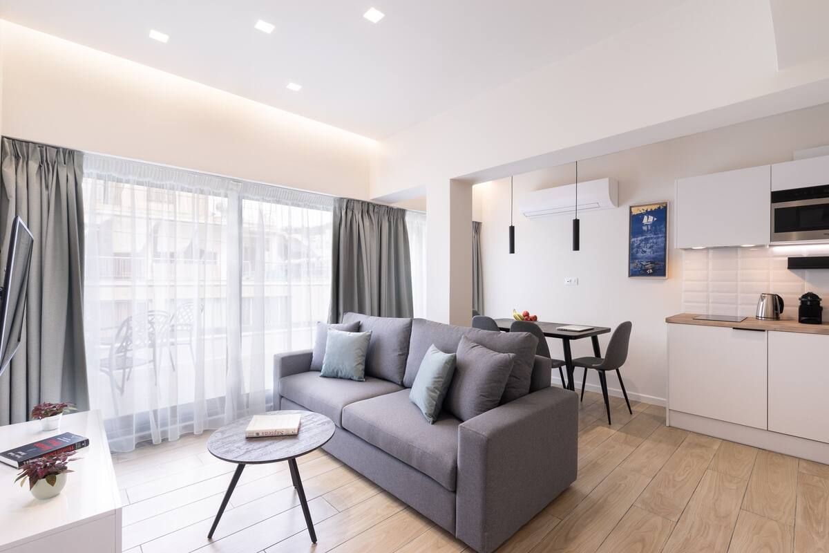 Una sala de estar moderna con un sofá gris, una mesita redonda y una cocina con armarios blancos y un microondas.