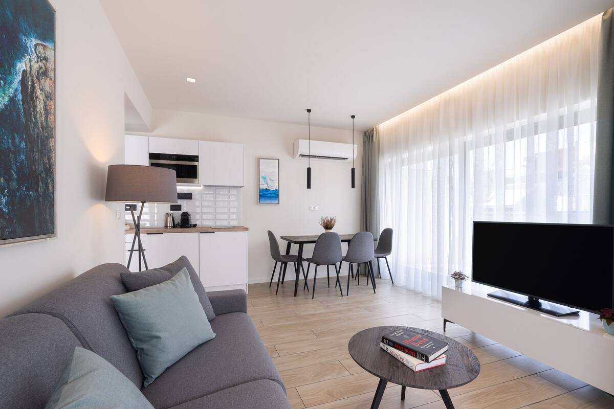 Una sala de estar moderna con un sofá gris, una mesa de centro con libros, un televisor de pantalla plana y una zona de comedor con una mesa y sillas.