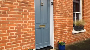 A red brick building with a blue door, a window with a plant, and two flower pots on the ground. - A Character Cottage in Historical Canterbury - Pass the Keys (Canterbury)