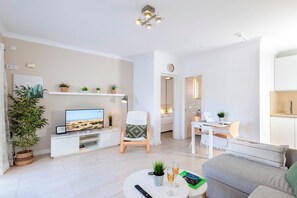A modern living room with a flat-screen TV, a sofa, a coffee table with drinks, and a dining area with a laptop and chair.