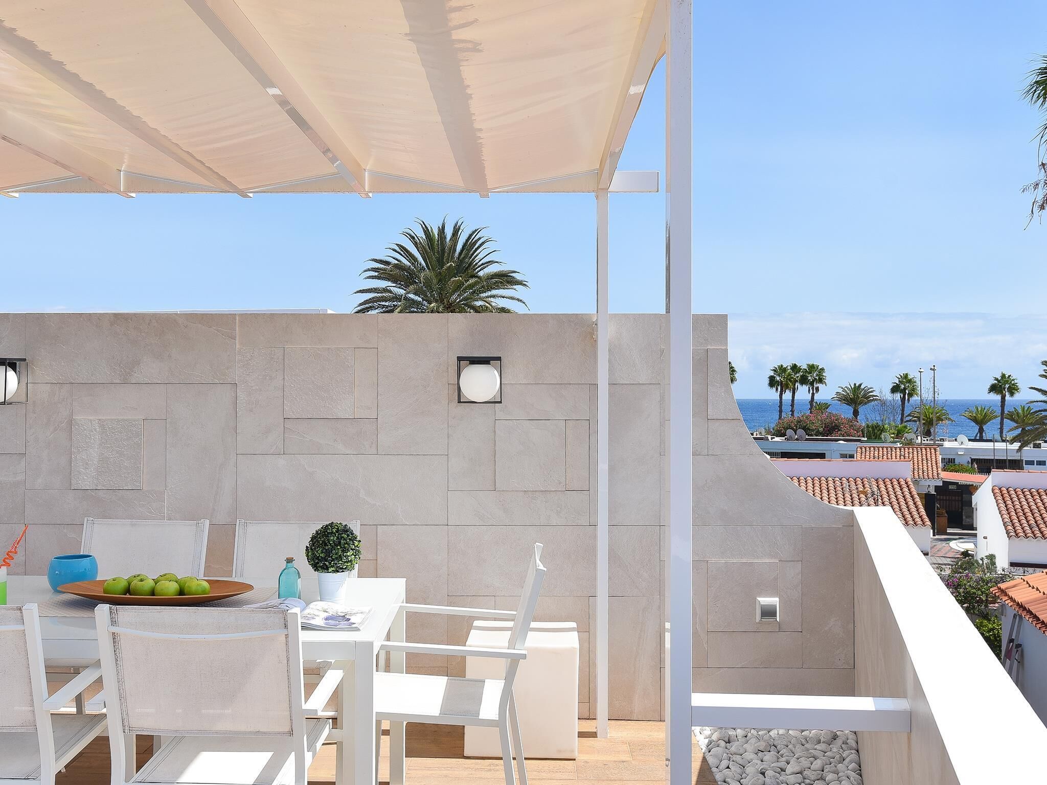 Una terraza con mesa y sillas, una planta en maceta y vistas al mar y palmeras.