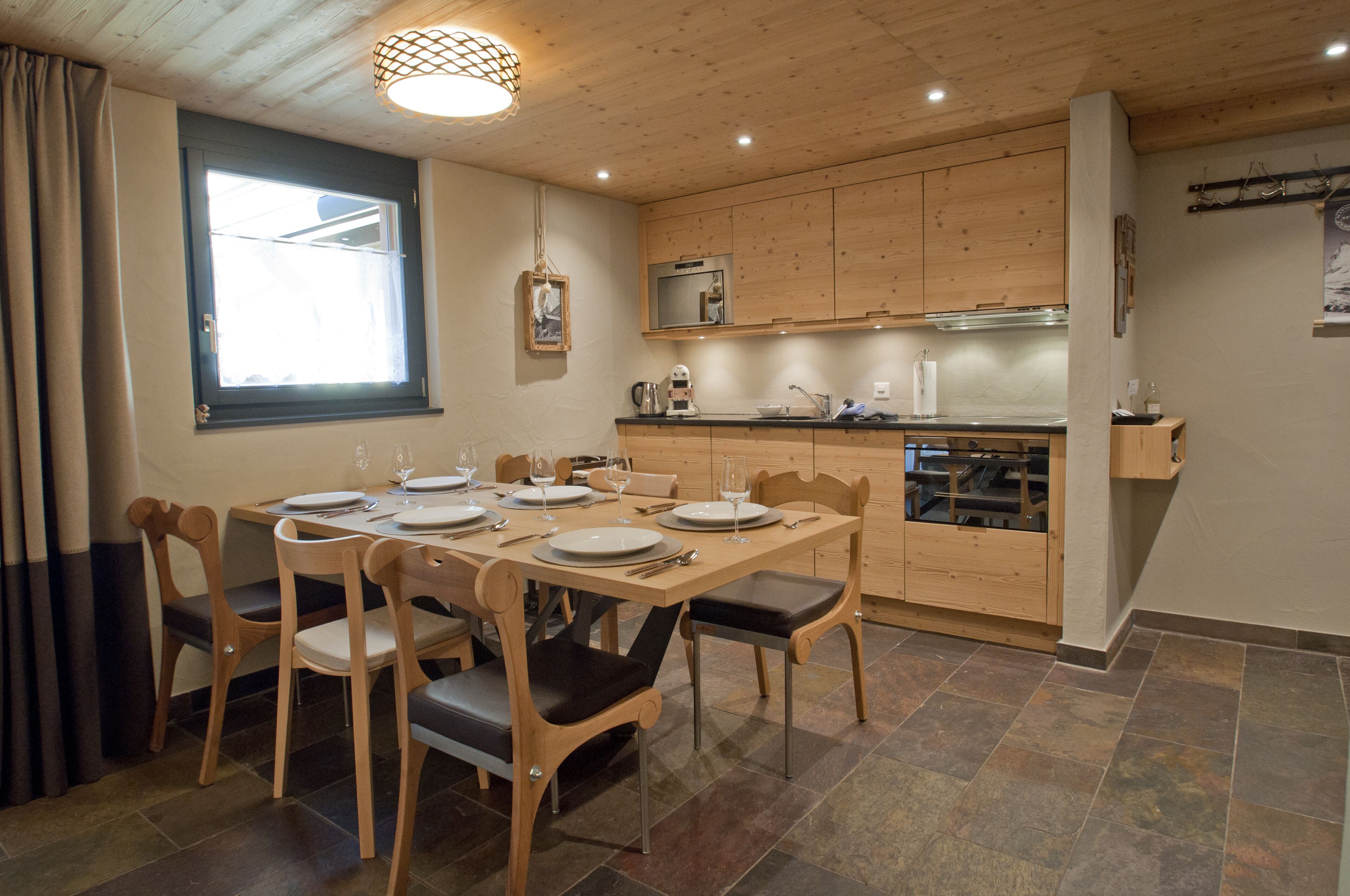 A dining area with a wooden table set for four, wooden chairs, a kitchen with wooden cabinets, and a window with curtains.