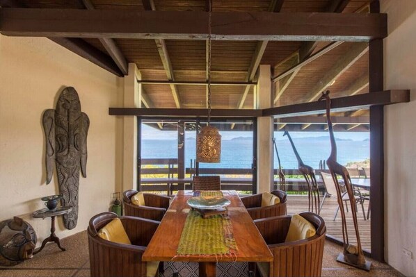 Enjoy meals in this spacious dining area with a long table and comfortable chairs, surrounded by a high ceiling and a large window offering a view of the ocean.
