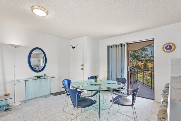 A modern room with a glass dining table, blue chairs, a round mirror, and a balcony with a grill.