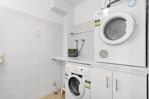 A laundry room with a front-loading washer and dryer, a small shelf with a bucket, and a shower area with a hose.