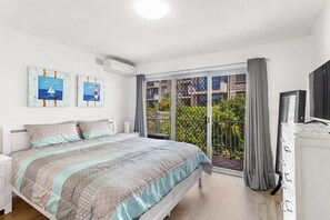 Een slaapkamer met een bed, een televisie en een balkon met uitzicht op gebouwen en groen.